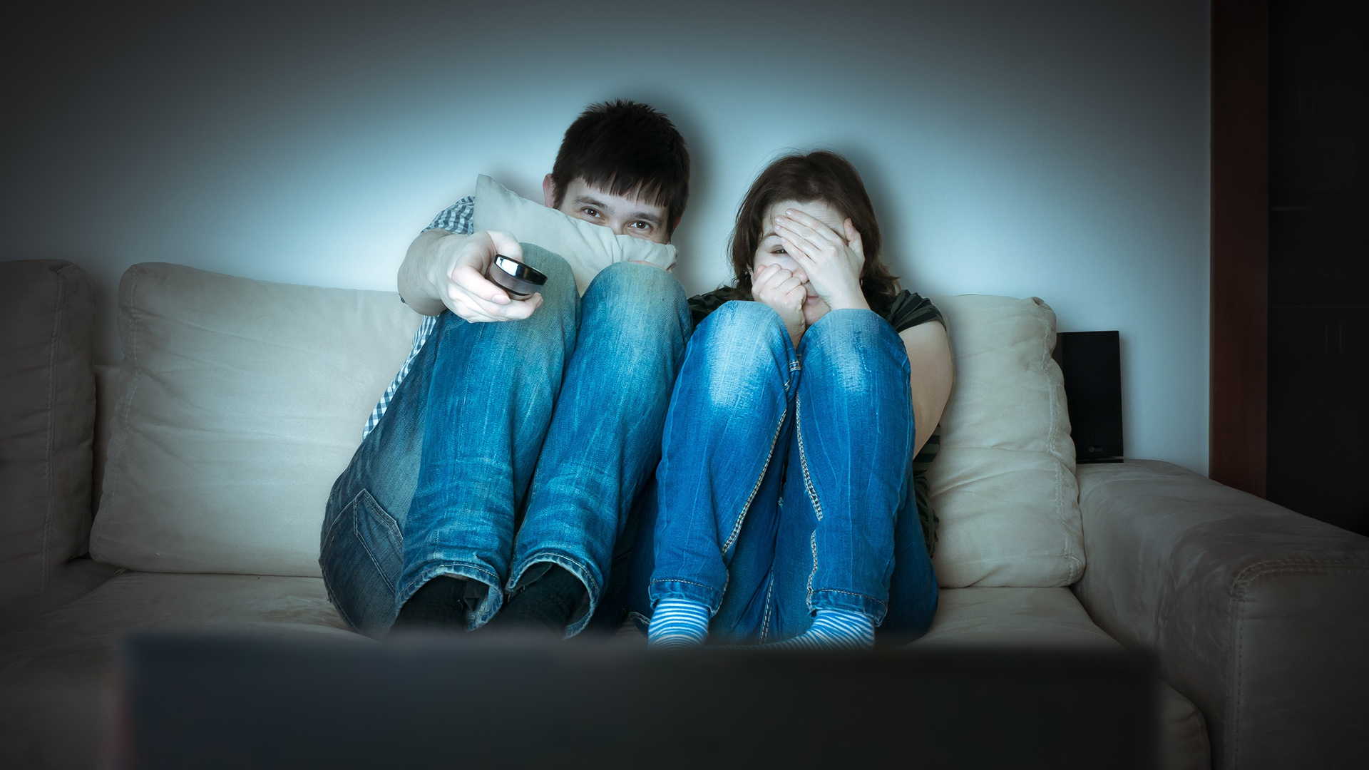 Photo of a couple on the couch, covering their eyes. They are watching a scary movie.